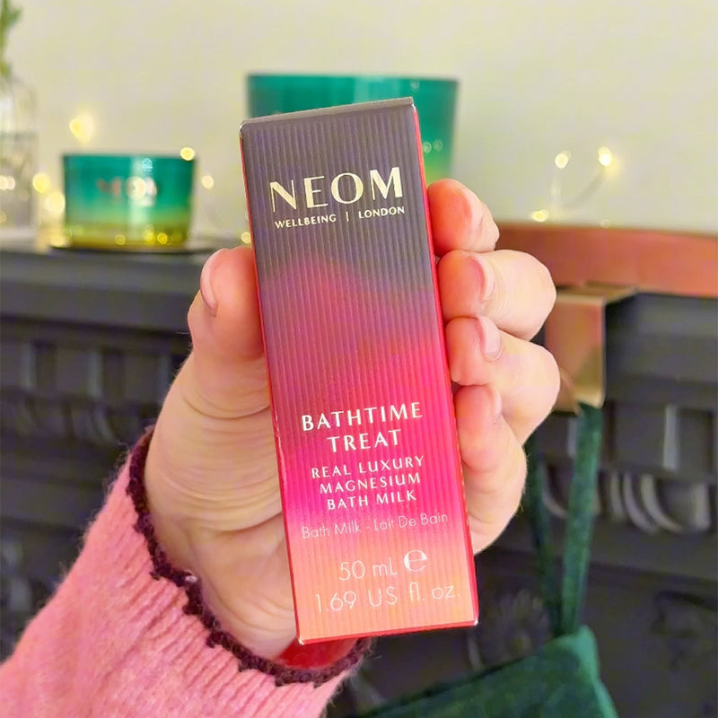 Hand holding NEOM Bathtime Treat Real Luxury Magnesium Bath Milk 50ml in front of candles on a fireplace.