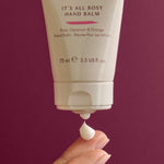 Hand balm tube labeled 'It's All Rosy' with a hand applying balm against a purple background