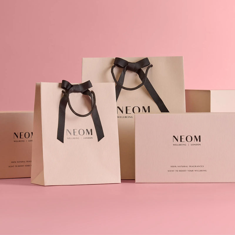 NEOM luxury gift boxes and ribbon-tied gift bags displayed on pink background.
