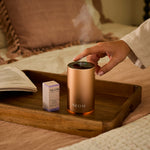 NEOM electric pod mini+ diffuser on a wooden tray with a hand interacting, surrounded by a cosy indoor setting.
